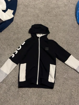 Svart och vit hoodie från BOSS - Snygg hoodie från BOSS i svart och vitt med dragkedja framtill och stor huva. Ärmarna har vita muddar och ena ärmen har BOSS tryckt i stora vita bokstäver. Två fickor framtill och cool kontrastdesign. Perfekt för en trendig streetwear-look.