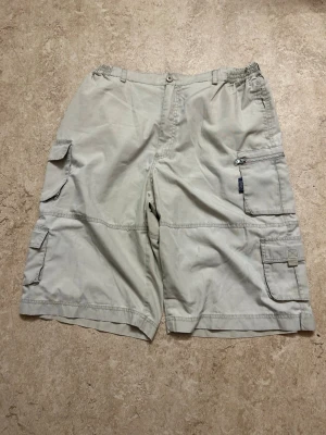 Beige cargoshorts med fickor - Säljer ett par beige cargoshorts med flera stora fickor på sidorna och baktill. Shortsen har dragkedja och knapp framtill samt elastisk midja för extra komfort. Perfekta för sommarens äventyr och har en avslappnad passform.