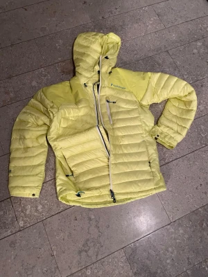 Gul dunjacka Peak Performance - Säljer en gul dunjacka från Peak Performance, modell HELI DOWN J. Jackan har huva, dragkedja framtill och en bröstficka med dragkedja samt innerficka. Tillverkad i lätt och smidig polyamid, perfekt för kalla dagar. Snygg quiltad design och justerbara detaljer vid ärmslut och nederkant.