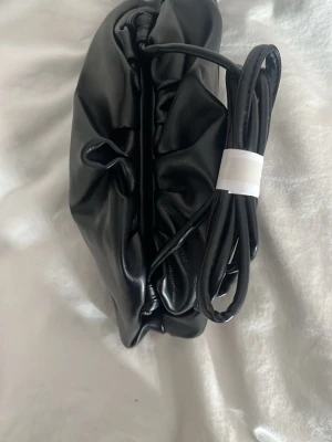 Svart Dumplingbag!  - ALDRIG ANVÄNT ! Använder inte pågrund av att jag råkade beställa två! Trendig svart axelväska med mjuk, puffig form och lång axelrem. Väskan har en minimalistisk design utan synliga detaljer och är tillverkad i slät skinnimitation. Perfekt för dig som gillar stilrena accessoarer.