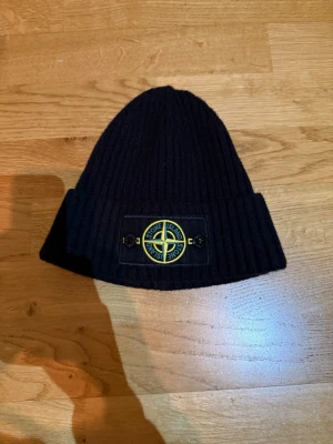 Svart ribbstickad mössa Stone Island - Svart ribbstickad mössa från Stone Island med uppvikt kant och den klassiska logotypen framtill. Märket sitter på en svart patch med gul och grön brodyr. Perfekt för dig som vill ha en stilren och trendig accessoar till hösten och vintern.