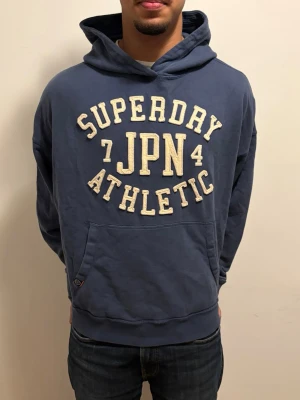 Extremt sällsynt vintage Blå Superdry hoodie  - Snygg blå hoodie från Superdry med stort broderat tryck i vitt och beige på bröstet där det står 'Superdry JPN Athletic'. Klassisk känguruficka framtill och huva med dragsko. Perfekt för en avslappnad och sportig stil. EXTREMT EFTERTRÄKTAD MODEL