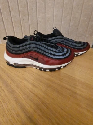 Nike Air Max 97 vinröd och svart - Säljer ett par Nike Air Max 97 sneakers i vinrött, svart och grått med vita detaljer. Skorna har den ikoniska vågiga designen, synlig Air-sula och snörning. Materialmix av syntet och textil. Perfekta för dig som vill ha en unik och sportig look.