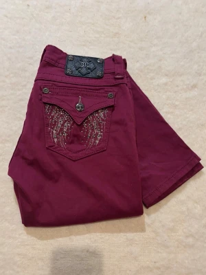 Vinröda skinny jeans från Miss Me - Snygga vinröda skinny jeans från Miss Me med coola detaljer och broderier på bakfickan. Jeansen har dekorativa nitar och knappar samt ett unikt mönster i silver på fickan. Materialet är en mix av bomull och elastan för skön stretch. Innerbenslängden är 80cm.