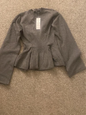 Mörkgrå peplumtopp i stickat material - Säljer en mörkgrå stickad peplumtopp från Gina. Toppen har lång ärm, hög hals och en markerad midja som ger en snygg siluett. Perfekt att styla med jeans eller kjol för en trendig look. Helt ny med prislapp