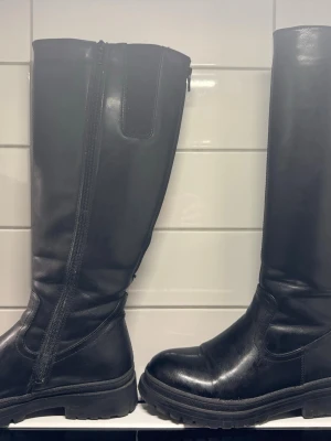 Svarta höga stövlar med dragkedja - Snygga svarta höga stövlar i skinnimitation med grov sula och dragkedja på insidan. Klassisk design med rund tå och platt sula, perfekta till höst- och vinterlooken. Passar till både jeans och kjol för en cool stil.