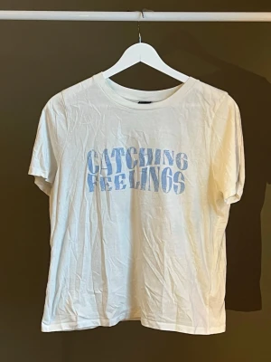 Vit t-shirt med blå text - Vit t-shirt med trycket 'CATCHING FEELINGS' i ljusblått framtill. Klassisk rund halsringning och korta ärmar. Tillverkad i mjukt bomullstyg för en avslappnad och bekväm stil. Perfekt för dig som gillar enkel men uttrycksfull design. Köpt från Gina Tricot, helt nyskick 🐚🪸