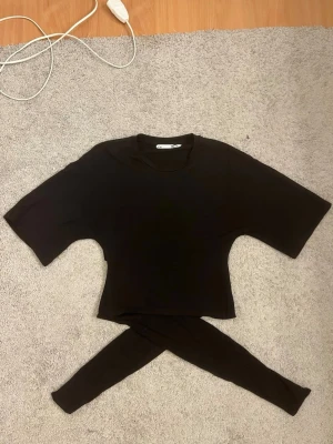 Svart croppad t-shirt med knyt från 157 - Svart croppad t-shirt från 157 med korta ärmar och rund hals. Baksidan har en snygg knyt-detalj som ger extra stilpoäng. Perfekt för dig som gillar enkel men trendig design. Mjuk och skön bomullskvalitet.