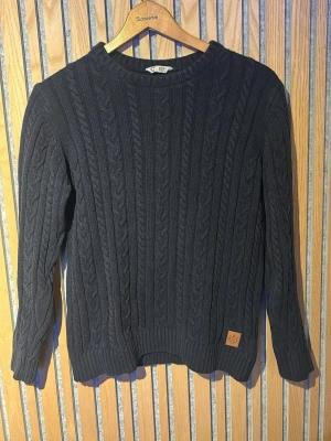 Mörkblå kabelstickad tröja Cubus - Snygg mörkblå kabelstickad tröja från Cubus med rund halsringning och ribbade muddar. Tröjan har ett klassiskt flätmönster och en liten läderpatch nertill. Perfekt för dig som gillar stilrena och tidlösa plagg.