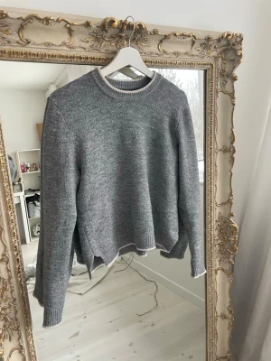 Grå stickad tröja med vit kant - Mysig grå stickad tröja med rund halsringning och vita detaljer vid hals, ärmslut och nederkant. Tröjan har en rak passform och är perfekt för lager-på-lager-stil. Enkel och stilren design som passar till det mesta.