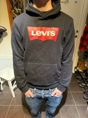 Levi's hoodie - Svart hoodie från Levi's med stor röd logga på bröstet. Klassisk känguruficka framtill och huva med dragsko. Mjuk bomullsblandning som är skön att ha på sig. Perfekt för en avslappnad och stilren look. 