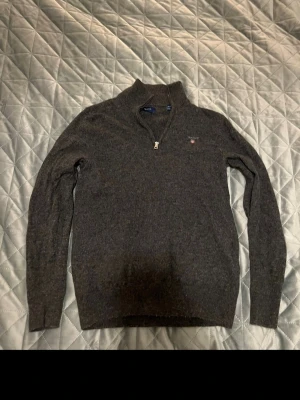 Mörkgrå halvzip tröja från Gant - Mörkgrå stickad tröja från Gant med halv dragkedja och liten broderad logga på bröstet. Tröjan har lång ärm och ribbade muddar vid ärmslut och nederkant. Perfekt för lager på lager och en stilren look.