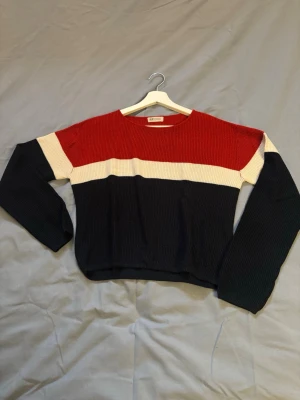 Randig stickad tröja från H&M - Säljer en snygg stickad tröja från H&M med breda blockränder i rött, vitt och mörkblått. Tröjan har rund halsringning och långa ärmar. Perfekt för dig som gillar en avslappnad men trendig stil.