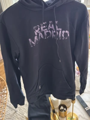 Svart Real Madrid hoodie - Snygg svart hoodie från Real Madrid med stor logga i vitt och lila på bröstet. Klassisk huva med dragsko och stor magficka framtill. Perfekt för dig som vill representera ditt favoritlag på ett stilrent sätt.