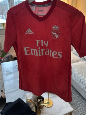Röd Real Madrid matchtröja Adidas - Snygg röd Real Madrid matchtröja från Adidas med Fly Emirates-tryck på bröstet och klubbmärke på vänster sida. Tillverkad i lätt och ventilerande material, perfekt för fotboll eller träning. Klassisk passform med korta ärmar och rund hals.