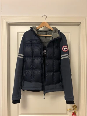 Vintage Canada Goose - Snygg blå dunjacka från Canada Goose med quiltad front, gråa detaljer och reflexränder på ärmarna. Jackan har huva, dragkedja och mjukt foder på insidan. Perfekt för kalla dagar och har en sportig look.