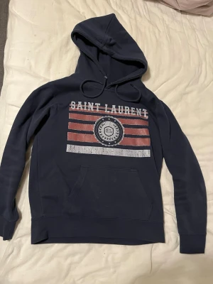 Mörkblå Saint Laurent hoodie - Mörkblå hoodie från Saint Laurent med stor huva och dragsko. Framsidan har ett tryck med texten 'SAINT LAURENT', röda och vita ränder samt ett emblem i mitten. Klassisk känguruficka framtill och ribbade muddar vid ärmar och nederkant. Perfekt för en chill och stilren look.