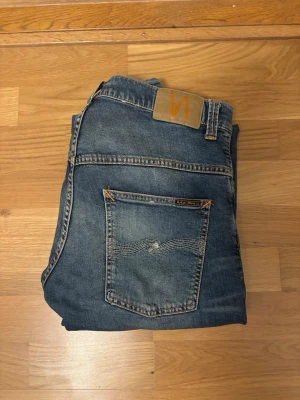 Nudies - Snygga blå jeans från Nudie Jeans med klassisk femficksdesign och läderpatch bak i midjan. Jeansen har raka ben och subtila slitningar framtill för en avslappnad look. Tillverkade i bomull med tydliga kontrastsömmar och signaturbroderi på bakfickan.