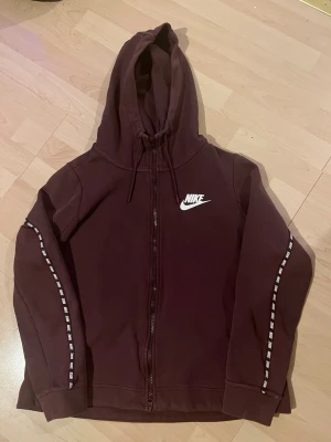 Vinröd Nike hoodie med dragkedja - Säljer en vinröd hoodie från Nike med dragkedja framtill och vit Nike-logga på bröstet. Hoodien har huva med dragsko och vita detaljer längs ärmarna. Perfekt för en avslappnad och sportig look. Materialet känns mjukt och bekvämt.
