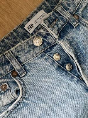 Ljusblå raka jeans från Zara - Säljer ett par ljusblå jeans från Zara med raka ben och klassisk femficksmodell. Jeansen har hög midja och knäppning med silverfärgade knappar. Materialet är 100% bomull för en skön och avslappnad känsla.