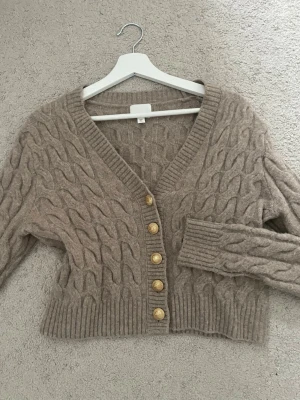 Beige kabelstickad kofta med guldknappar - Säljer en beige kabelstickad kofta med djup v-ringning och fem dekorativa guldknappar framtill. Koftan har ribbade muddar och är lite croppad i modellen. Perfekt för dig som gillar en mysig men ändå trendig look.