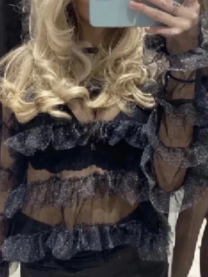 Svart glittrig volangblus - ❗️pris kan diskuteras❗️Säljer en transparent svart blus med glittriga volanger och lång ärm. Blusen har flera lager volangdetaljer och är gjord i ett tunt, syntetiskt material som ger en festlig look. Perfekt för dig som vill sticka ut med en unik topp! 