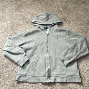 Grå hoodie från Polo Ralph Lauren - Klassisk grå hoodie från Polo Ralph Lauren med dragkedja framtill och huva. Liten broderad logga på bröstet och två fickor fram. Tillverkad i mjuk bomullsmix, perfekt för en avslappnad stil.