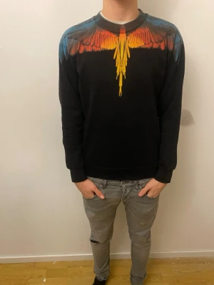 Svart Marcelo Burlon tröja med fågelmotiv - Säljer en svart sweatshirt från Marcelo Burlon med ikoniskt färgstarkt fågelmotiv över axlar och bröst i rött, orange, gult och blått. Tröjan har rund halsringning och ribbade muddar. Perfekt statement-plagg för dig som vill sticka ut.