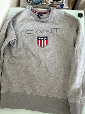 Grå sweatshirt från GANT - Snygg grå sweatshirt från GANT med broderad logga och sköld på bröstet. Klassisk rund halsringning och långa ärmar. Perfekt till vardags för en avslappnad men stilren look.