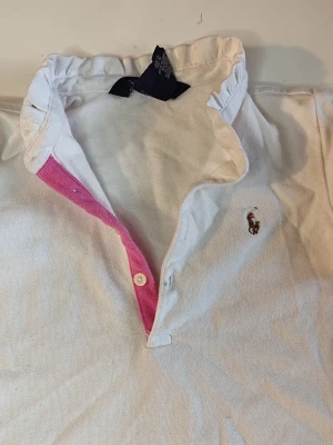 Vit pikétröja från Ralph Lauren - Klassisk vit pikétröja från Ralph Lauren med ikoniska broderade loggan på bröstet. Tröjan har krage, knäppning med två knappar och en snygg rosa detalj på insidan av knappslån. Perfekt till jeans eller kjol för en sportig look.  Du är varmt välkommen att kika på mina andra annonser.