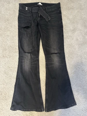 Svarta bootcut jeans från Gina Tricot - Säljer ett par svarta bootcut jeans från Gina Tricot i storlek L. Jeansen har slitningar vid knäna och en cool, utsvängd passform. Klassisk femficksmodell med knapp och dragkedja framtill. Perfekta för dig som gillar en edgy stil.