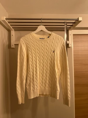 Vit kabelstickad tröja från Gant - Kabelstickad tröja i offwhite från Gant med rund halsringning och ribbade muddar. Klassisk design med diskret Gant-logo broderad på bröstet. Perfekt för dig som gillar stilrena och tidlösa plagg i garderoben.