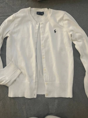 Vit kofta från Ralph Lauren - Klassisk vit kofta från Ralph Lauren med rund halsringning och knäppning framtill. Tröjan har ribbade muddar vid ärmslut och nederkant samt den ikoniska mörkblå logotypen broderad på bröstet. Perfekt att slänga över en t-shirt eller linne.