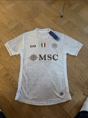 Napoli tröja - Bilderna visar en vit fotbollströja från Napoli med subtila mönster, bruna loggor och detaljer, samt texten 'Proud to be Napoli' på ryggen. 