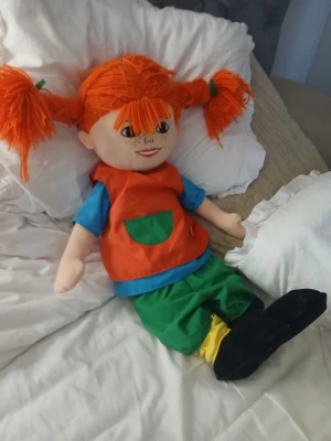 Stor Pippi nalle - Rolig och färgglad Pippi nalle med stora orange garnflätor, blå tröja, röd väst med grön ficka, gröna byxor och svarta skor. Nallen är mjuk och passar perfekt som lekkompis eller dekoration i rummet.