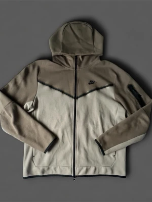 Nike tech fleece - Nike vindjacka i beige och brunt med hel dragkedja framtill och svart Nike-logga på br soppen. Jackan har huva, långa ärmar och en extra ficka med dragkedja på ena ärmen. Tillverkad i mjukt material, perfekt för en sportig look.