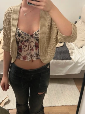 Beige kortstickad trekvartsärmad kofta - Säljer en beige, kortstickad kofta med öppen front och trekvartsärmar. Koftan har ett luftigt, grovstickat mönster och breda ribbade kanter. Perfekt att slänga över ett linne eller topp för en avslappnad look.