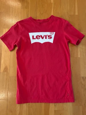 Röd Levi's t-shirt med logga - Säljer en klassisk röd t-shirt från Levi's med den ikoniska vita loggan tryckt på bröstet. T-shirten har rund hals och korta ärmar, perfekt för en avslappnad och stilren look. Tillverkad i mjuk bomull för skön komfort.