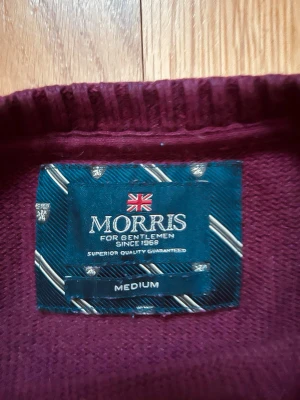 Vinröd stickad tröja från Morris - Snygg vinröd stickad tröja från Morris med rund halsringning och vit broderad MORRIS-text på bröstet. Tröjan har ribbade muddar vid ärmslut och nederkant samt en klassisk passform.
