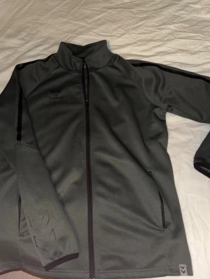 Mörkgrön träningsjacka från Hummel - Snygg mörkgrön träningsjacka från Hummel med hel dragkedja och svarta detaljer längs ärmarna. Jackan har två sidofickor med dragkedja och Hummels logga på bröstet samt de klassiska pilarna på ärmen. Tillverkad i ett mjukt och stretchigt material.