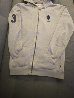 Ljusgrå hoodie från USPA med dragkedja - Ljusgrå hoodie från USPA med dragkedja framtill, huva och fickor. Mörkblå USPA-logga på bröstet och siffra 3 på ärmen. Mjuk insida och ribbade muddar. Perfekt för en avslappnad och sportig stil.