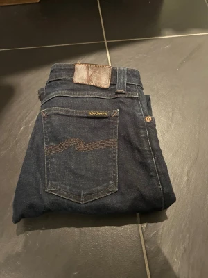 Mörkblå jeans från Nudie Jeans - Snygga mörkblå jeans från Nudie Jeans med klassisk femficksdesign, slitningar och subtila söm-detaljer på bakfickan. Jeansen har en rak passform och är tillverkade i slitstarkt bomullsdenim. Perfekta för dig som gillar stilrena och tidlösa jeans.