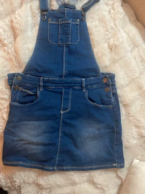 Blå jeanskjol med hängslen - Säljer en blå jeanskjol med hängslen och fickor fram och bak. Kjolen har klassiska jeansdetaljer som knappar, bälteshällor och kontrastsömmar. Perfekt för dig som gillar en avslappnad och trendig stil. Storleken står 12 år - för 152 cm långa tjejer - men kan användas för de som har xs och s i storlek då den är strechig 