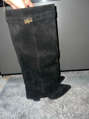 Svarta mockastövlar med metallspänne - Snygga svarta stövlar i mocka med spetsig tå och klack. Stövlarna har en dekorativ metallspänne upptill som ger en exklusiv känsla. Perfekta att styla till kjol eller klänning för en trendig look. Aldrig använda