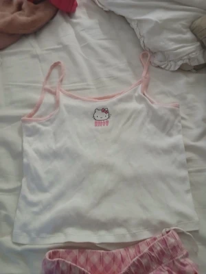 Hello Kitty pyjamas rosa/vit - Sött pyjamas från Hello Kitty med ett vitt linne med rosa kantband och tryck framtill. (OBS, Det är inte ett set, byxorna går att köpa seperat men notera att det är olika stotlek på byxorna och linnet)