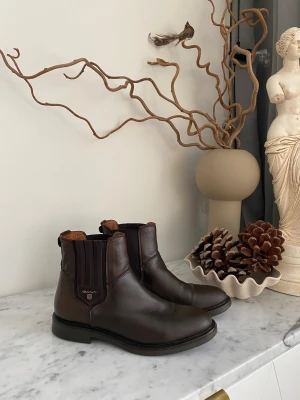Bruna Chelsea boots från Gant - Säljer ett par klassiska Chelsea boots från GANT i storlek 39. Använda men i mycket fint skick, med ett stilrent uttryck som håller år efter år. Det här är bootsen som funkar överallt – kontoret, middagen, AW eller när du vill vara välklädd utan att det känns stelt.  Tidlös design, bra passform och den där självklara GANT-kvaliteten som bara blir bättre med tiden.  Här får du ett par kvalitetsboots för cirka en fjärdedel av nypriset.  Pris: 550 kr  Hämtas i Sundsvall eller skickas mot frakt. Spa