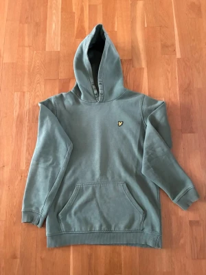 Grön hoodie från Lyle & Scott Junior - Snygg grön hoodie från Lyle & Scott Junior med klassisk huva och känguruficka framtill. Diskret logga i gult och svart på bröstet. Tillverkad i mjukt bomullsmaterial som är skönt att ha på sig. Perfekt för en avslappnad och trendig look.