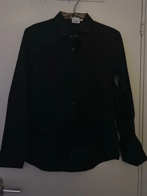 Svart klassisk skjorta från Burberry - Stilren svart skjorta från Burberry med klassisk krage och knäppning framtill. Skjortan är helt ny och aldrig använd.