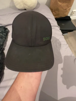 Svart keps från BOSS - Snygg svart keps från BOSS med klassisk böjd skärm och broderad grön BOSS-logga framtill. Kepsen har en stilren design och är tillverkad i ett slitstarkt tygmaterial. Perfekt för dig som vill ha en enkel men trendig accessoar.