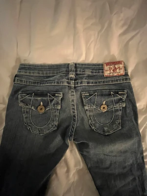 Ljusblå True Religion Jeans - Säljer ett par ljusblå jeans från True Religion med kontrastsömmar och stora fickor bak med dekorativa knappar. Jeansen har låg midja och lite bootcut men mer åt det raka hållet. Säljer då jag inte använder dem längre💕 Kom privat för mer bilder eller frågor!